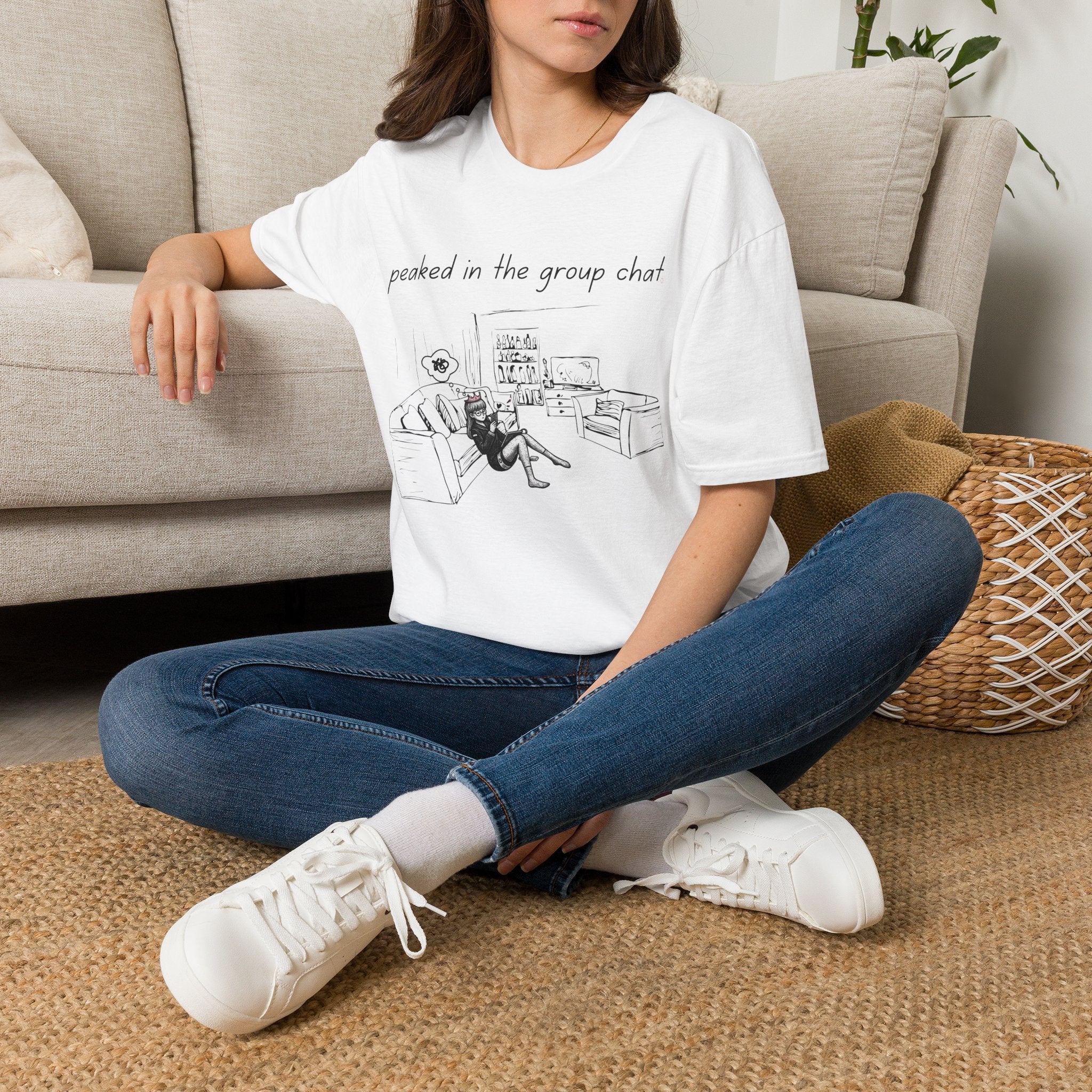 White tee with 'peaked in the group chat' in minimalist type above a simple line-sketch living room with couch and phone — front view
