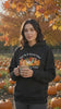A young woman wearing the black hoodie stands in a sunlit pumpkin patch in late afternoon. The camera frames her from the waist up, the hoodie's painted pumpkin harvest illustration centered and clearly visible on her chest. She holds a warm drink in both hands. Golden autumn light filters through orange and amber maple trees behind her, and a few leaves drift down slowly. The camera makes one slow, gentle push-in